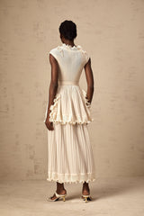 A woman wearing a long white sleeveless maxi dress with ruffle details and pleats standing against a plain background