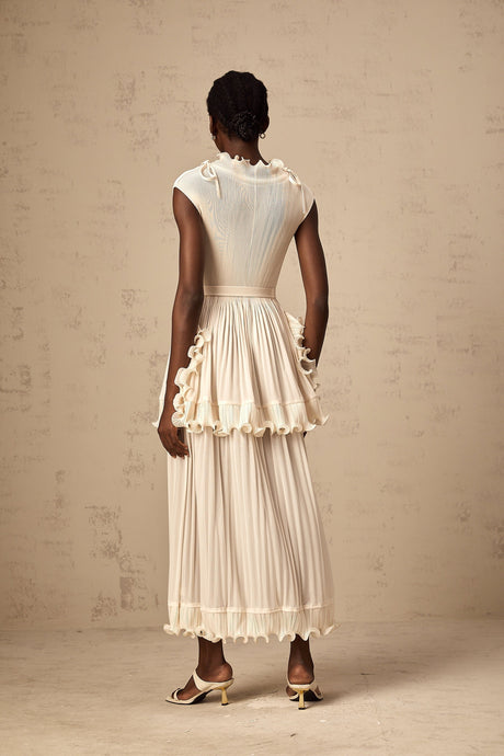 A woman wearing a long white sleeveless maxi dress with ruffle details and pleats standing against a plain background
