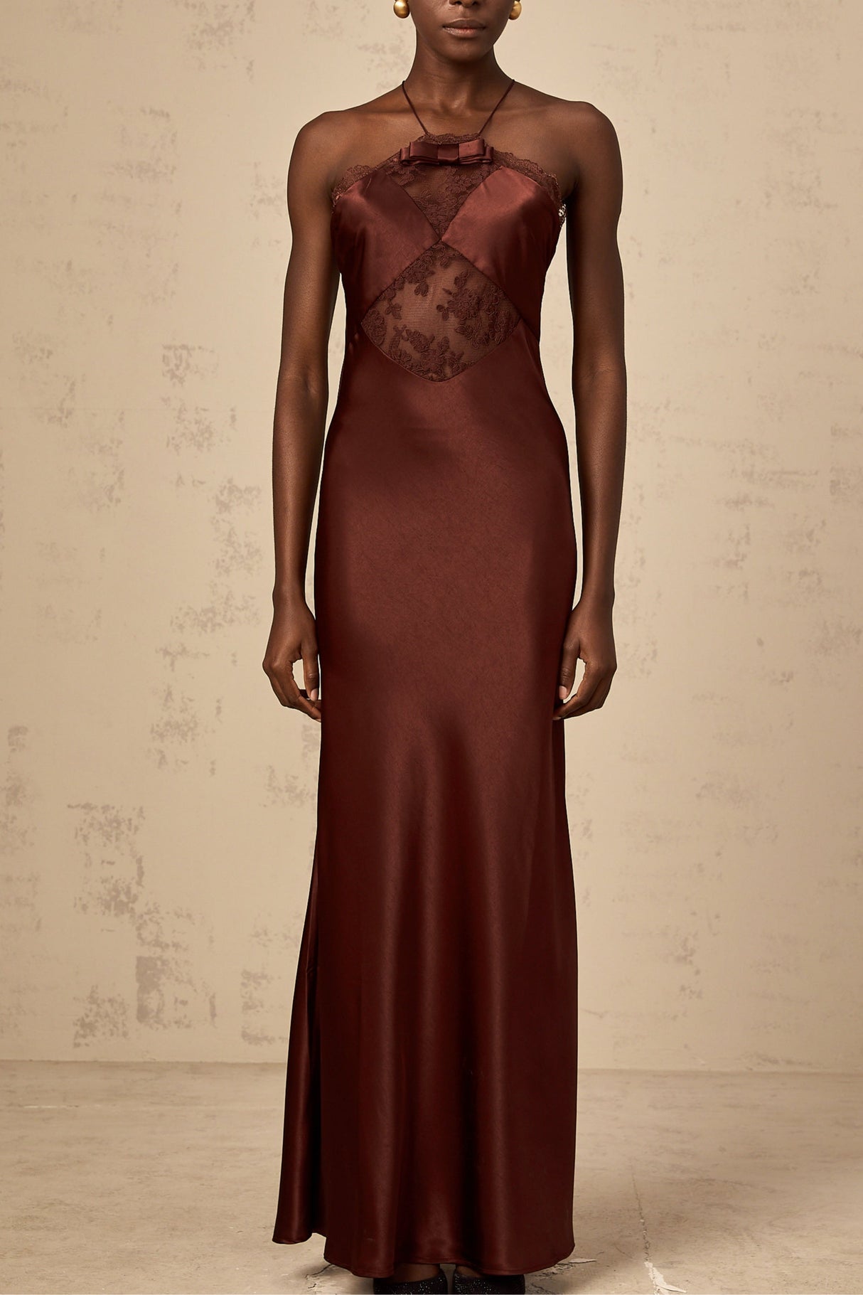 A woman wearing a long winered maxi dress with lace panels and satin fabric standing elegantly against a plain background