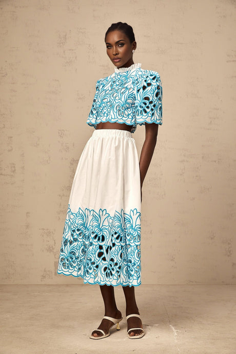 A woman wearing a matching blue twopiece outfit consisting of a sleeveless crop top with cutout details and embroidery paired with a high waisted skirt featuring similar embroidery and cutout designs