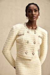 A woman wearing a matching knit cardigan and skirt set in a light color with embellished details The cardigan has long sleeves and the skirt falls to knee length Both pieces feature decorative embroidery or beading