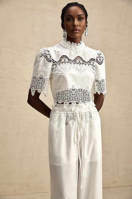 A woman wearing a matching set consisting of a white lace trim cropped top with cutouts and white trousers The top has short sleeves and the trousers are high waisted Both pieces feature delicate lace details