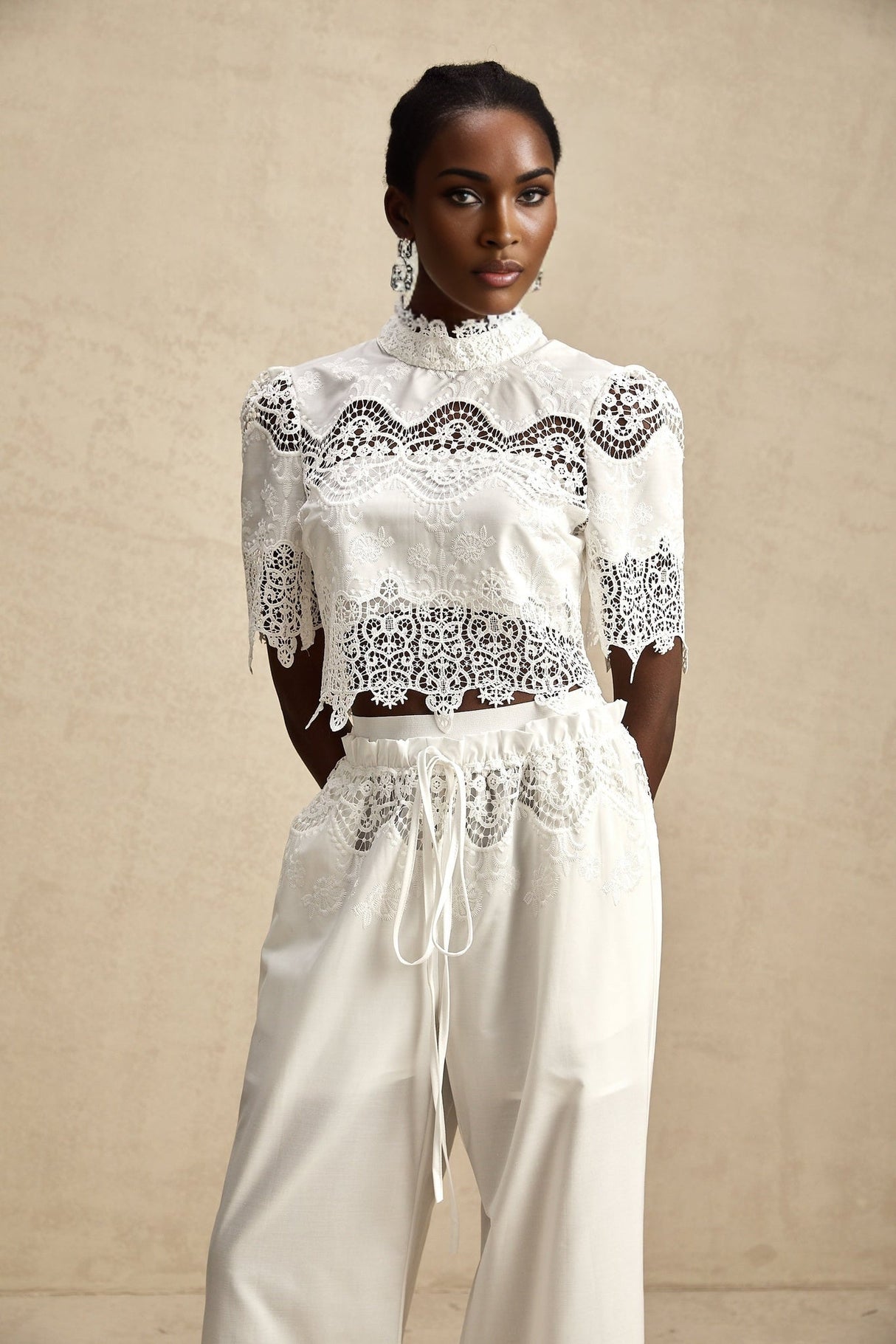 A woman wearing a matching two piece outfit consisting of a cropped top and trousers in light fabric The top features lace trim and cutout details while the trousers are high waisted Both pieces are in a coordinating color and style