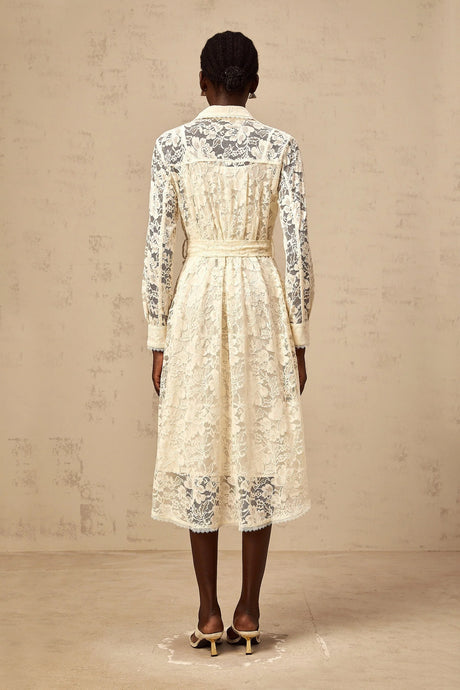 A woman wearing a neutralcolored midi length shirt dress with guipure lace detailing standing in a relaxed pose