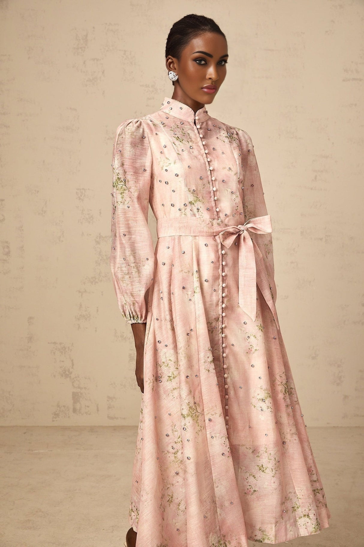 A woman wearing a pink midi dress with floral print and embellishments standing against a plain background
