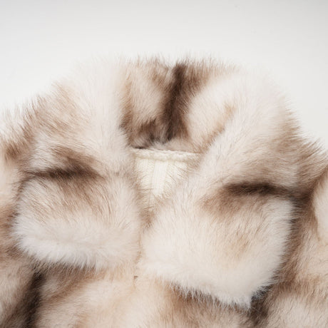 A woman wearing a plush beige faux fur jacket with ribbed knit sleeves and collar standing against a plain background