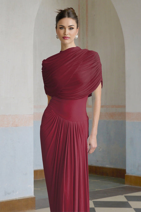 A woman wearing a red asymmetric pleated maxi dress with a flowing silhouette standing against a plain background
