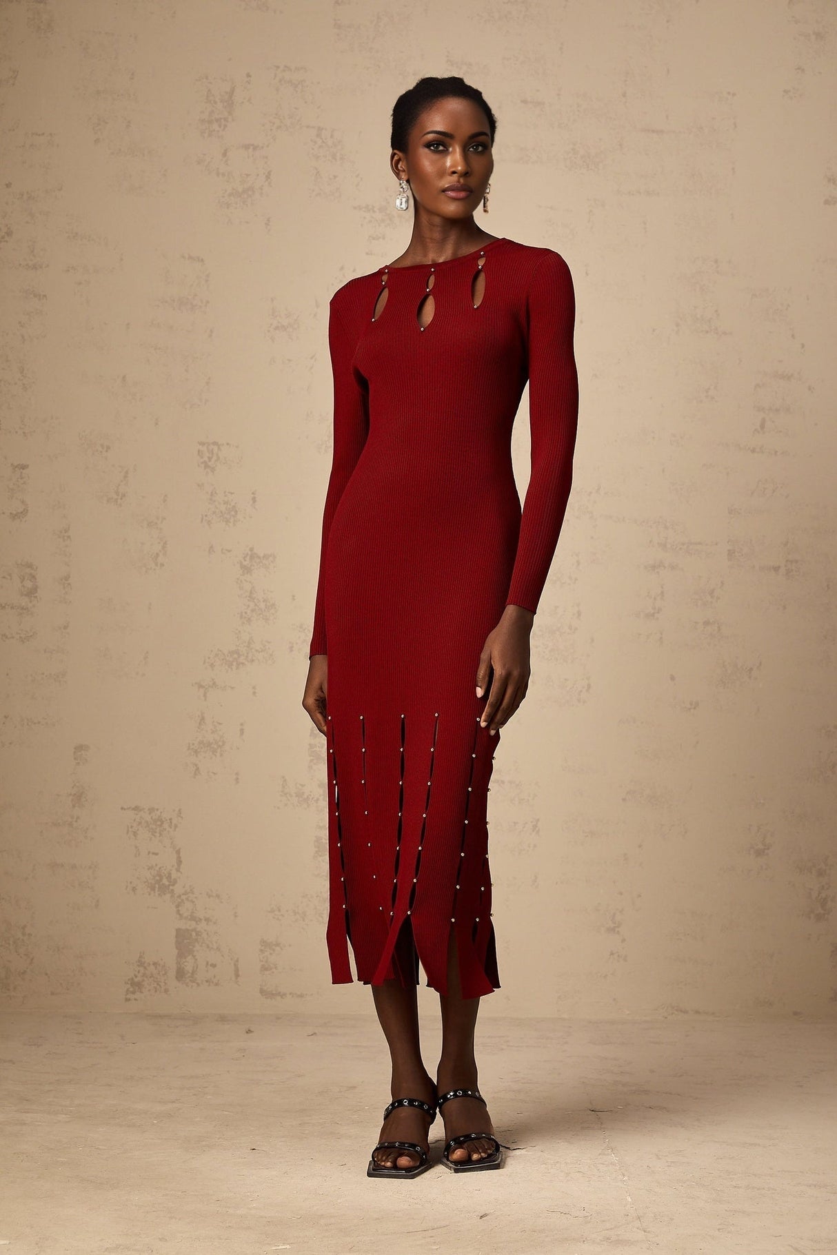 A woman wearing a red knitted midi dress with long sleeves and beaded details The dress features cutout sections and falls to mid calf length
