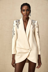 A woman wearing a shimmering blazer dress covered in sequins with embellished details standing against a plain background