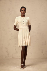 A woman wearing a short beige knitted dress with pleated skirt and delicate pointelle trim standing in a neutral background