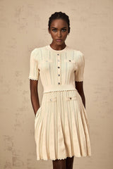 A woman wearing a short beige knitted dress with pleats and delicate pointelle trim details standing in a neutral setting