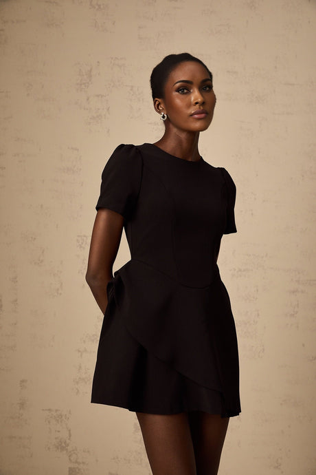 A woman wearing a short black dress with an asymmetrical neckline and ruffled detailing standing against a plain background