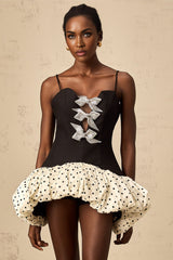A woman wearing a short colorful dress with polka dots and a large bow accent at the waist The dress features contrasting color blocks and has a playful retro inspired style
