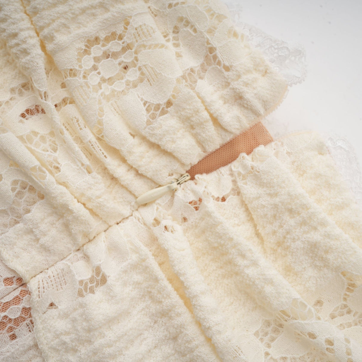 A woman wearing a short neutral colored floral lace mini dress with flared cuffs standing against a plain background