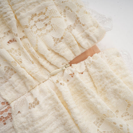 A woman wearing a short neutral colored floral lace mini dress with flared cuffs standing against a plain background