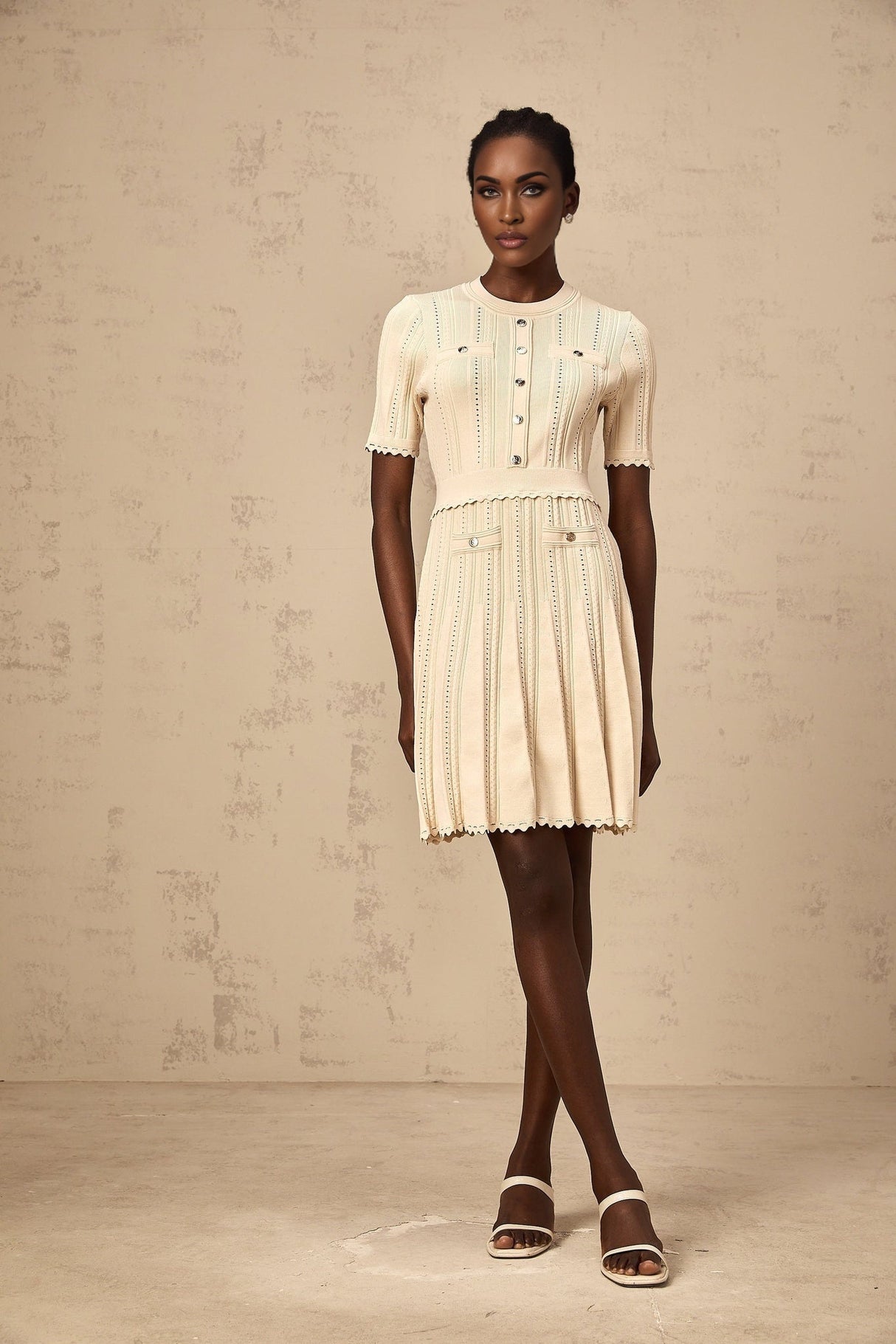 A woman wearing a short neutral colored knit dress with pleated skirt and delicate pointelle trim detailing