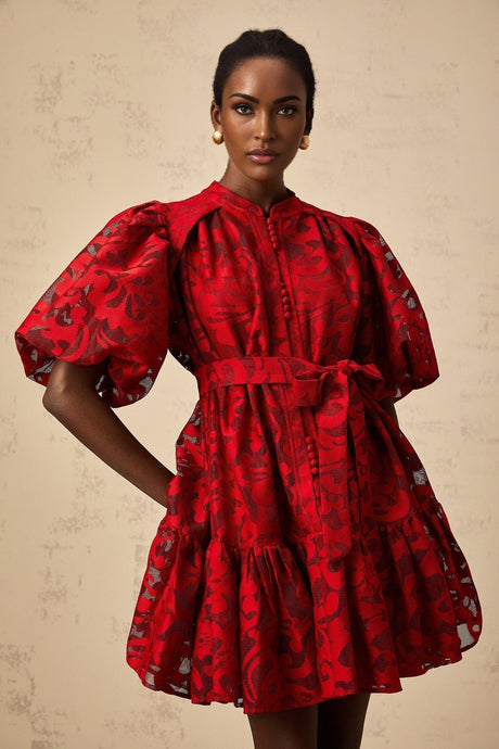A woman wearing a short red jacquard dress with a semi sheer design standing in a relaxed pose
