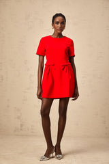 A woman wearing a short red mini dress with short sleeves and a bow detail at the front standing against a plain background
