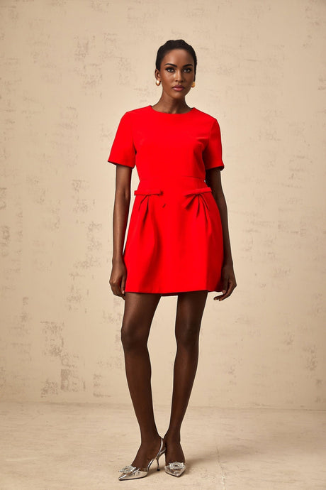 A woman wearing a short red mini dress with short sleeves and a bow detail at the front standing against a plain background