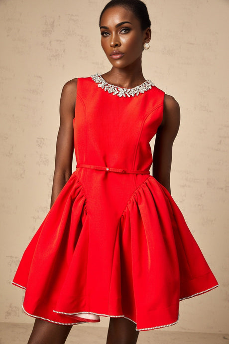 A woman wearing a short red pleated dress with embellishments standing against a plain background