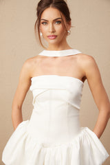 A woman wearing a short white halterneck dress with ruffled details standing against a plain background
