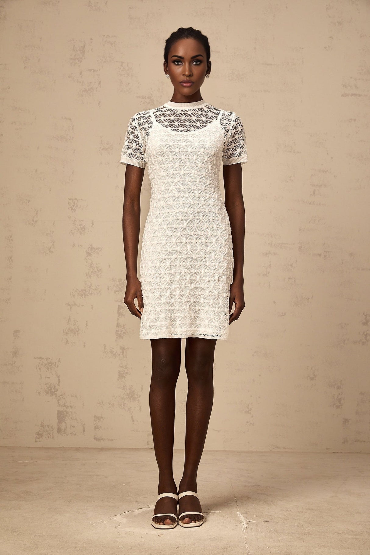 A woman wearing a short white semisheer knit dress standing in a neutral background