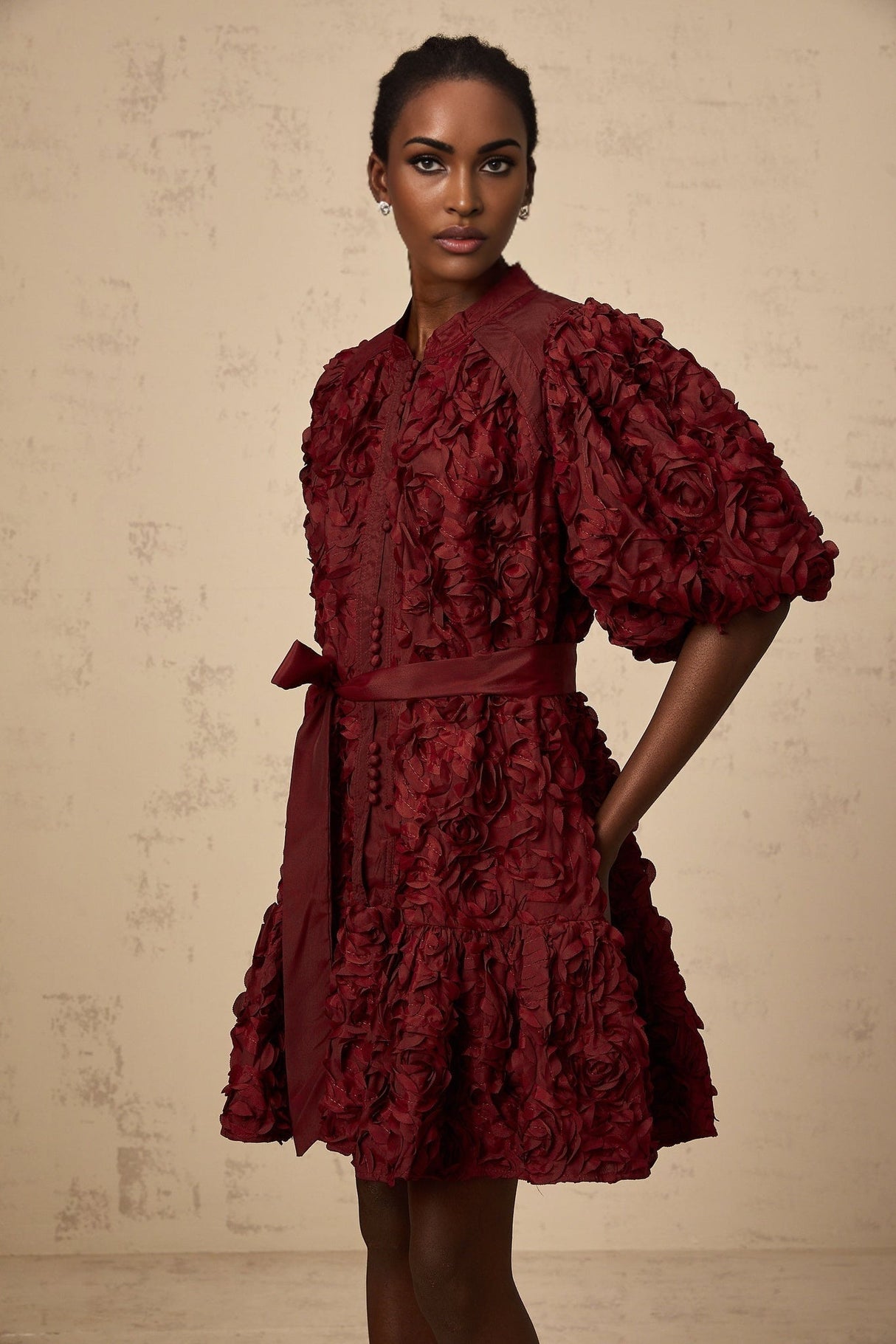 A woman wearing a short winered dress with three dimensional petallike puffs decorating the fabric standing against a plain background