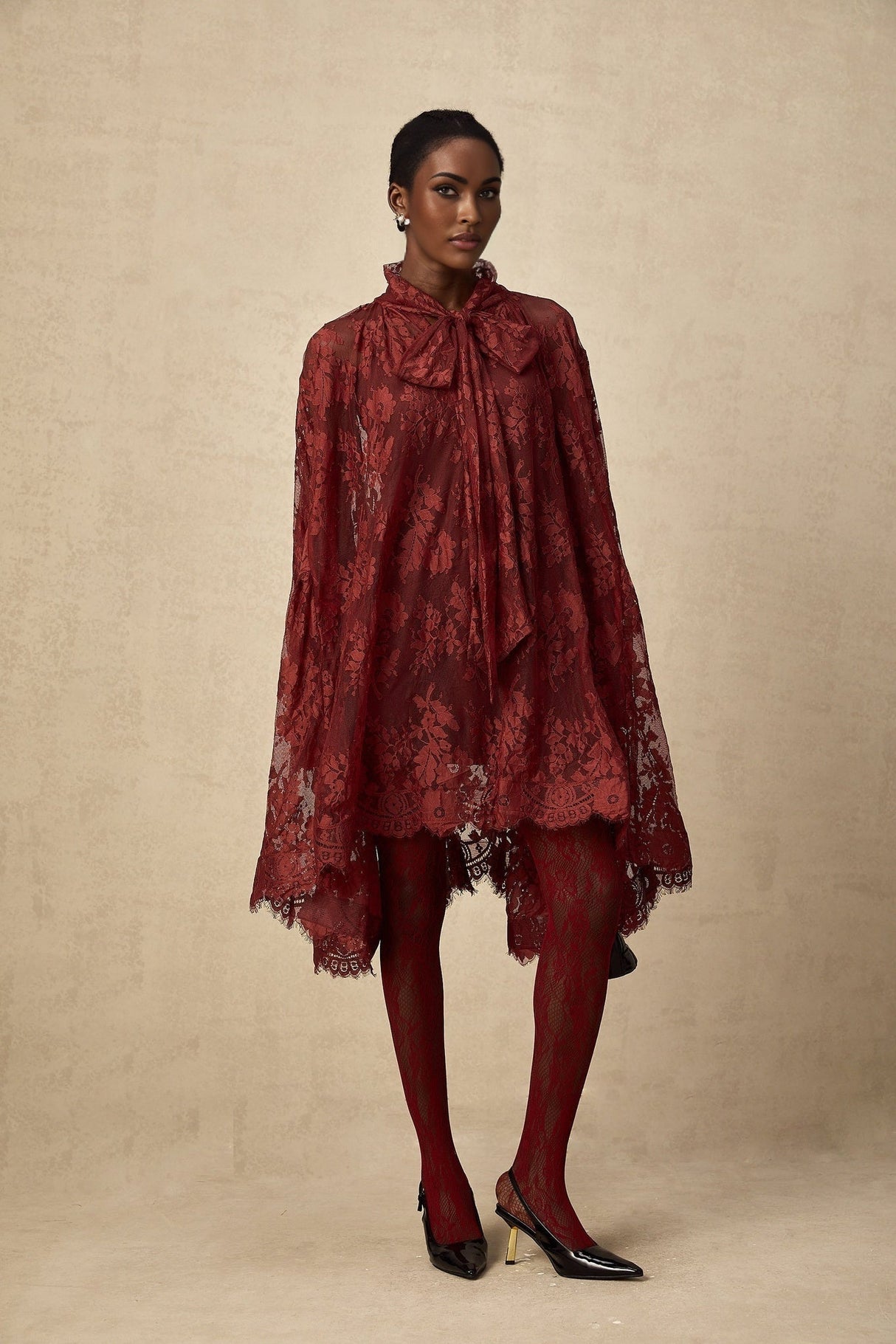 A woman wearing a vibrant red oversize dress made of delicate chantilly lace standing in a relaxed pose against a plain background