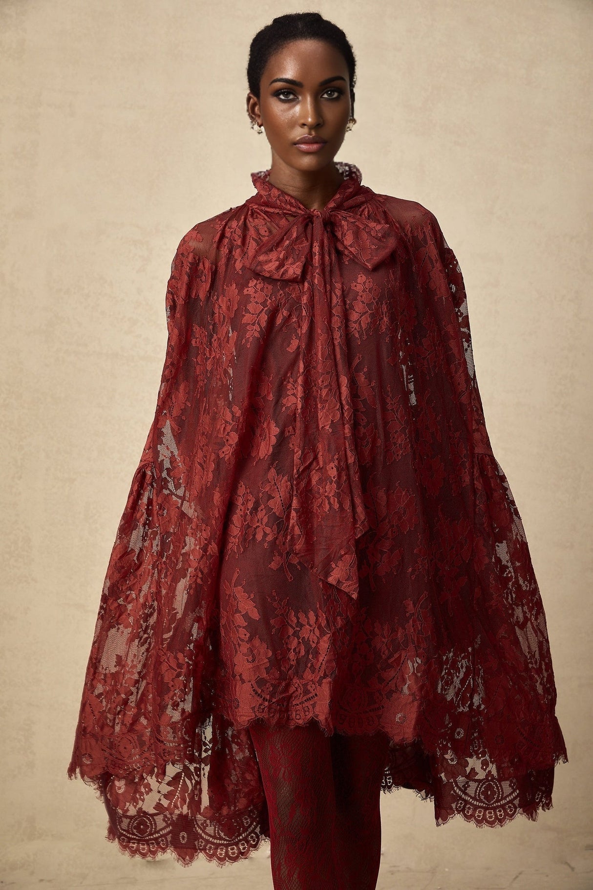 A woman wearing a vibrant red oversize dress made of delicate chantilly lace standing in a relaxed pose against a plain background