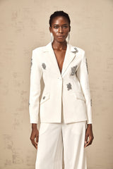 A woman wearing a white blazer and matching trousers embellished with crystal details standing confidently against a plain background