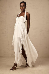 A woman wearing a white chiffon maxi dress with ruffles and a halter neck design standing against a plain background