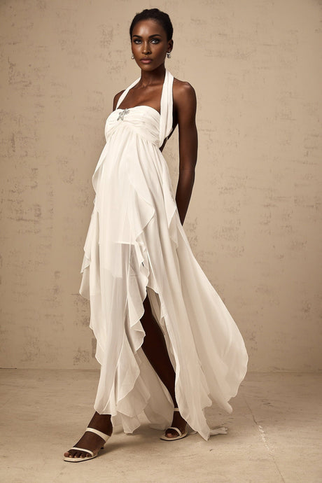 A woman wearing a white chiffon maxi dress with ruffles and a halter neck design standing against a plain background