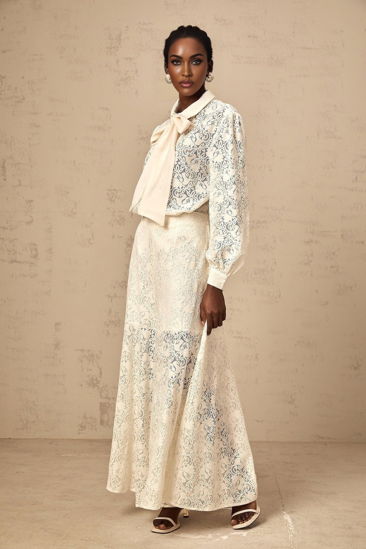 A woman wearing a white guipure lace matching set consisting of a shirt and trousers standing against a plain background