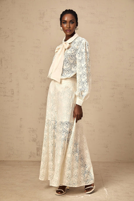 A woman wearing a white guipure lace matching set consisting of a shirt and trousers standing against a plain background