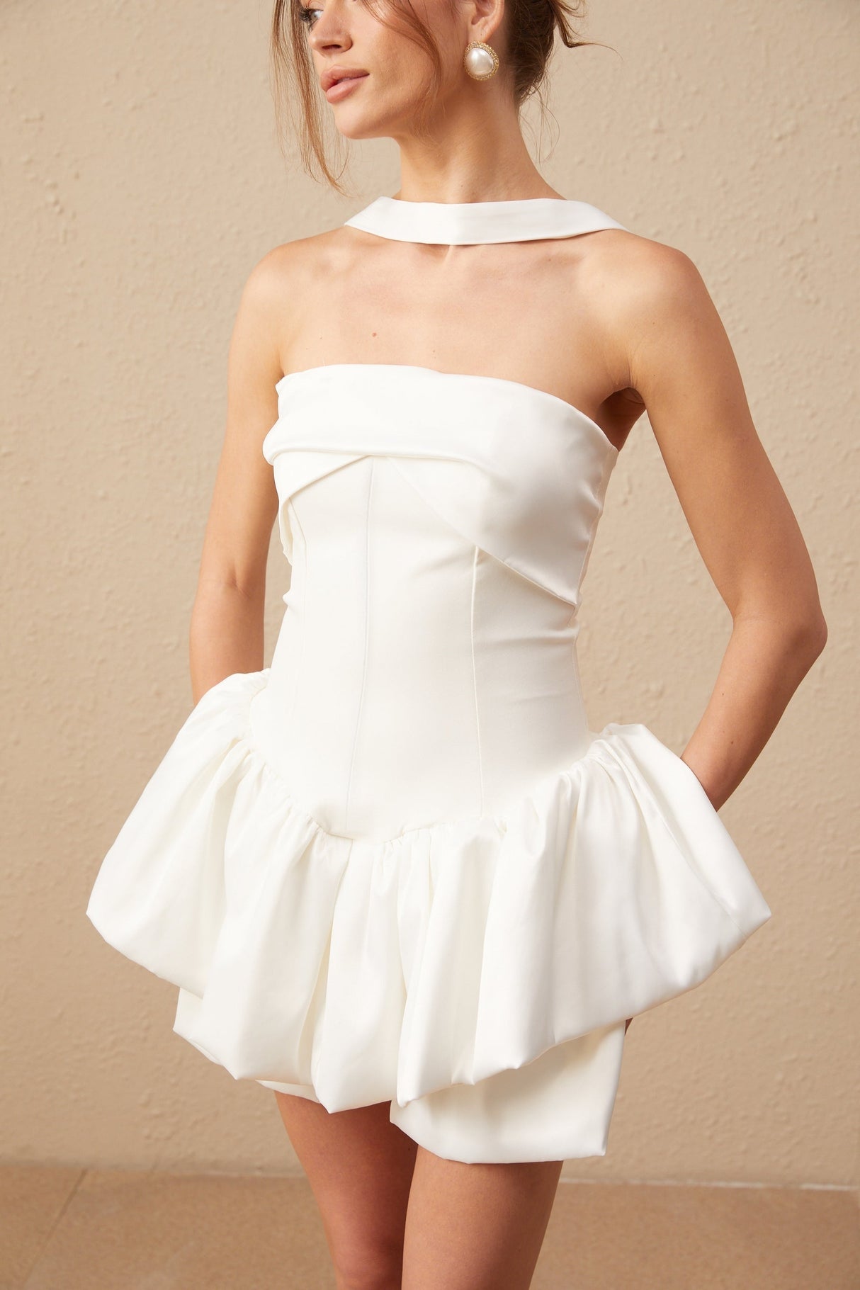 A woman wearing a white halterneck mini dress with ruffled details standing against a plain background