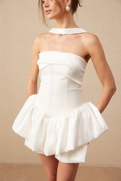 A woman wearing a white halterneck mini dress with ruffled details standing against a plain background