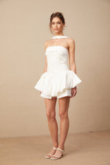 A woman wearing a white halterneck mini dress with ruffled details standing in a relaxed pose against a plain background