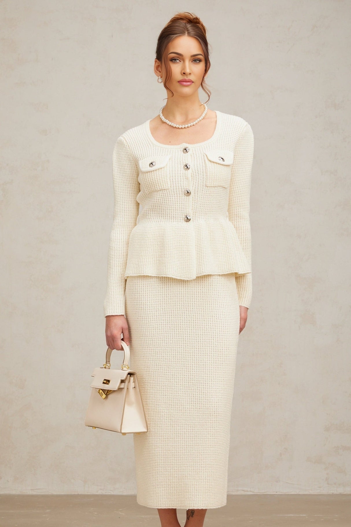 A woman wearing a white knitted matching set consisting of a sleeveless top and a knee length skirt standing against a plain background