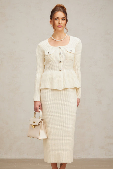 A woman wearing a white knitted matching set consisting of a sleeveless top and a knee length skirt standing against a plain background