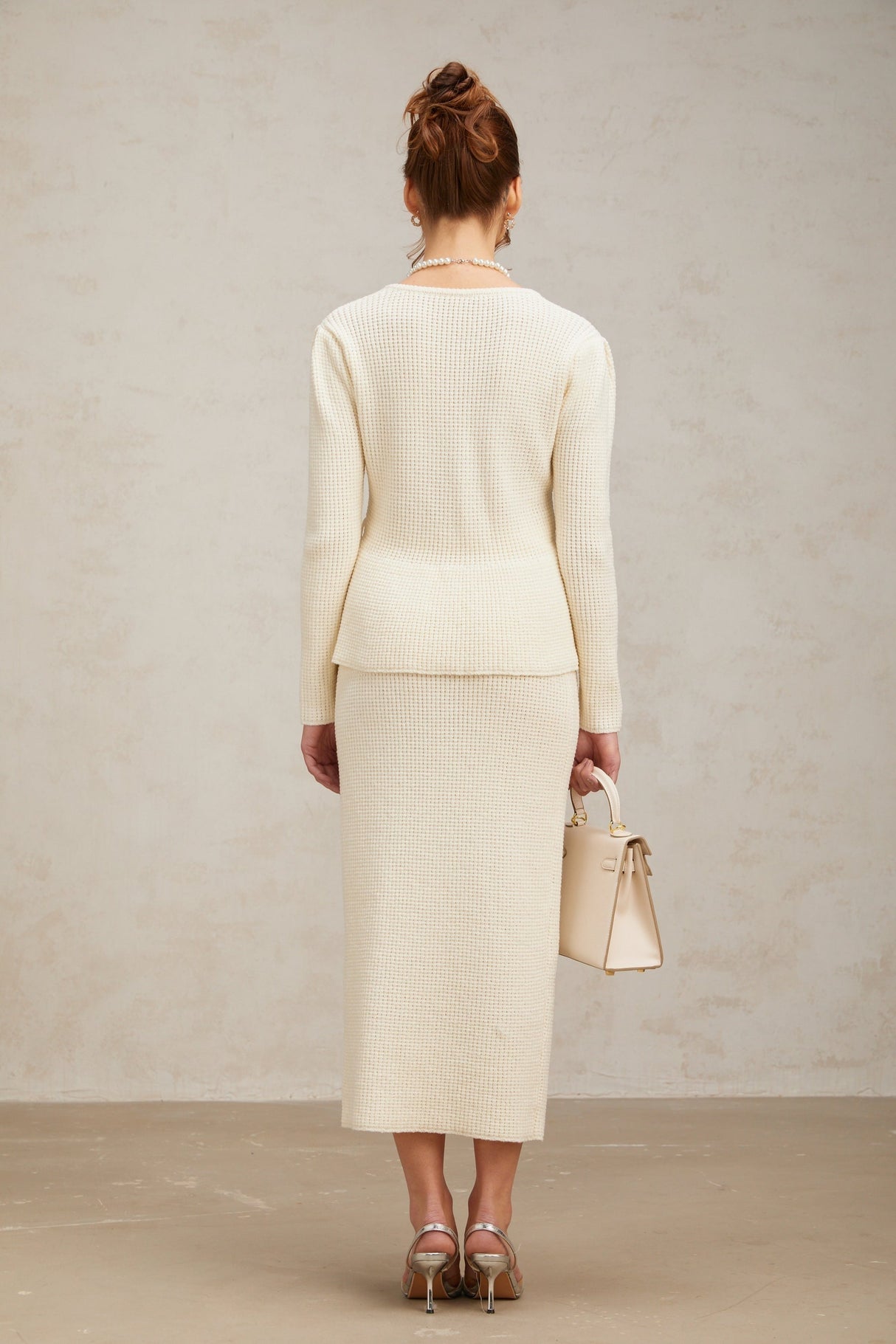 A woman wearing a white knitted matching set consisting of a sleeveless top and a knee length skirt standing against a plain background