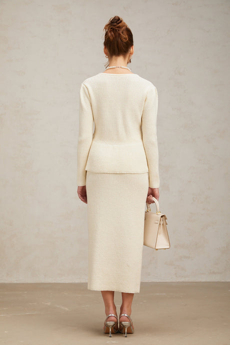 A woman wearing a white knitted matching set consisting of a sleeveless top and a knee length skirt standing against a plain background
