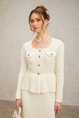 A woman wearing a white knitted two piece outfit consisting of a sleeveless top and matching mini skirt standing against a plain background