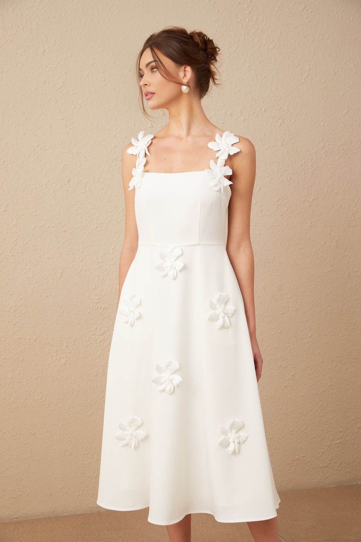 A woman wearing a white midi dress with a square neckline adorned with faux flower details standing against a plain background