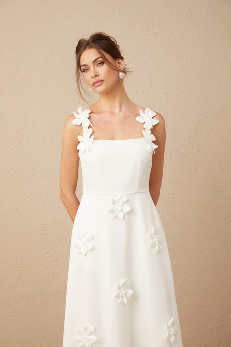 A woman wearing a white midi dress with a square neckline adorned with faux flower embellishments standing against a plain background