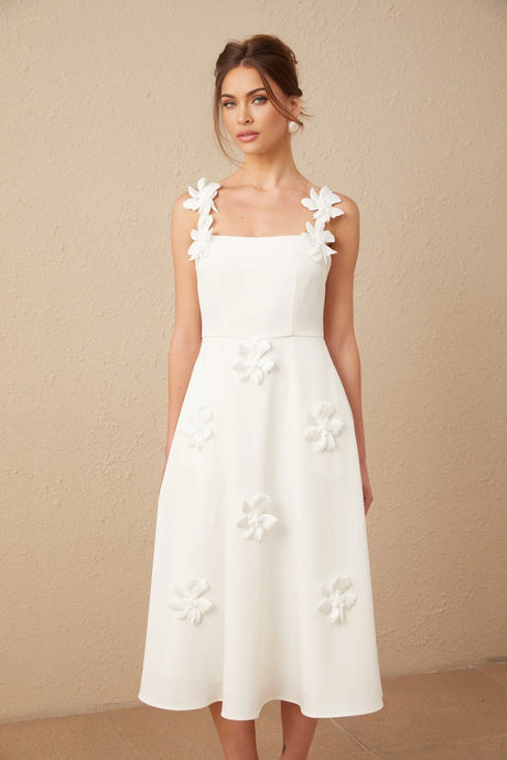 A woman wearing a white midi dress with a square neckline and faux flower details standing in a neutral setting