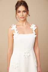 A woman wearing a white midi dress with a square neckline and faux flower embellishments standing in a neutral background
