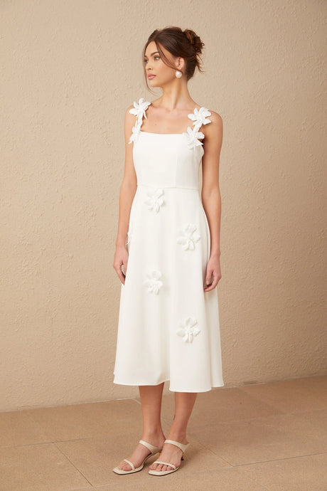 A woman wearing a white midi dress with a square neckline and faux flower embellishments standing in a neutral setting