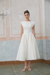 A woman wearing a white midi dress with cap sleeves and an open back design standing in a relaxed pose