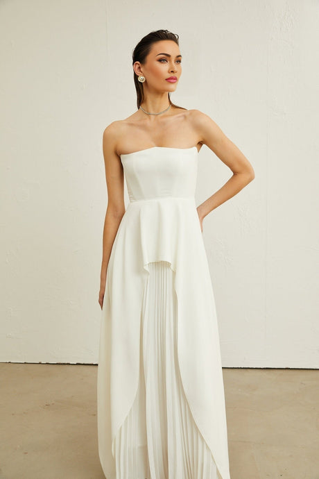 A woman wearing a white offshoulder maxi dress with pleated details standing against a plain background