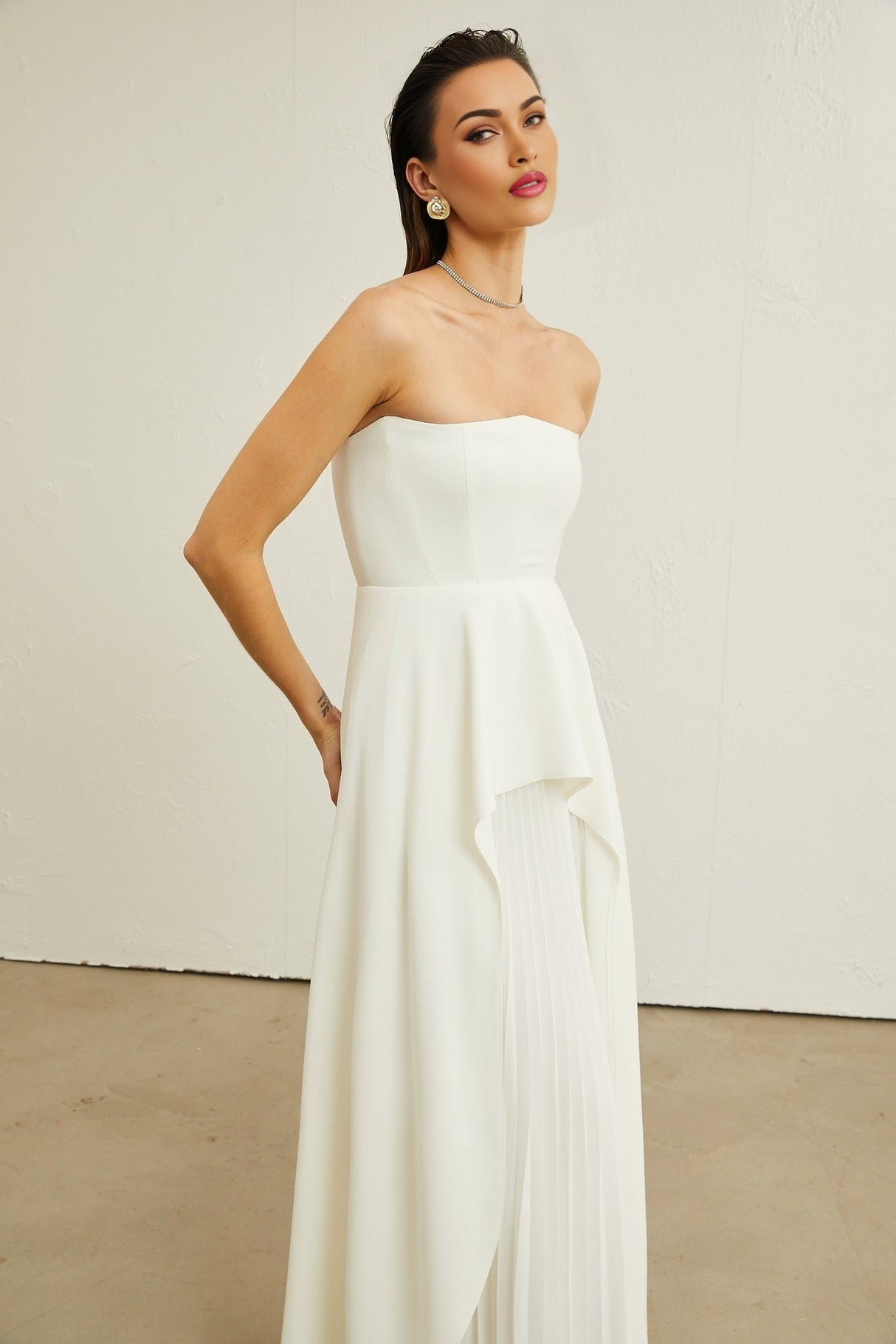 A woman wearing a white offshoulder maxi dress with pleated details standing against a plain background